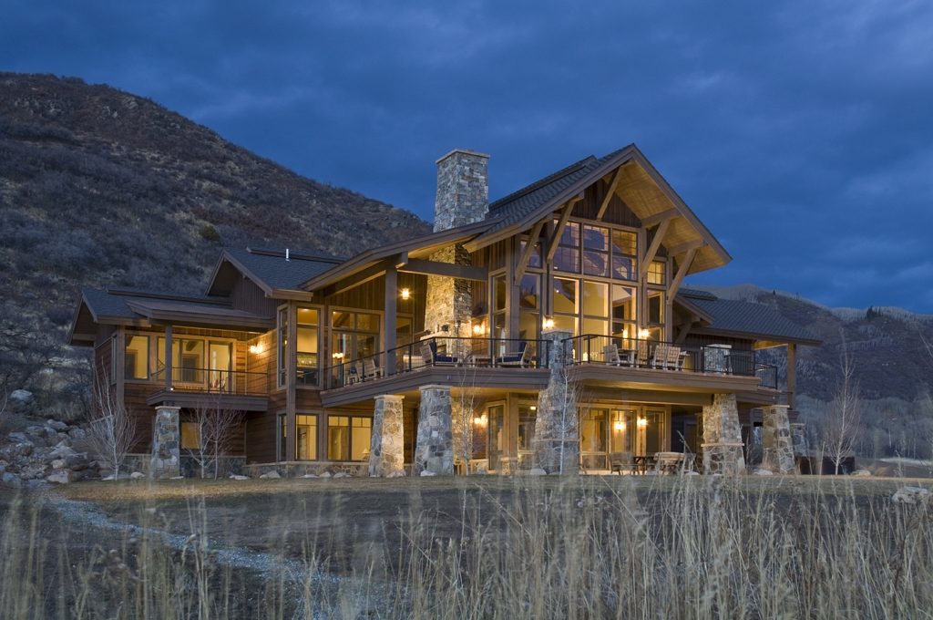Custom timber and stone home glowing at dusk beside a mountain meadow